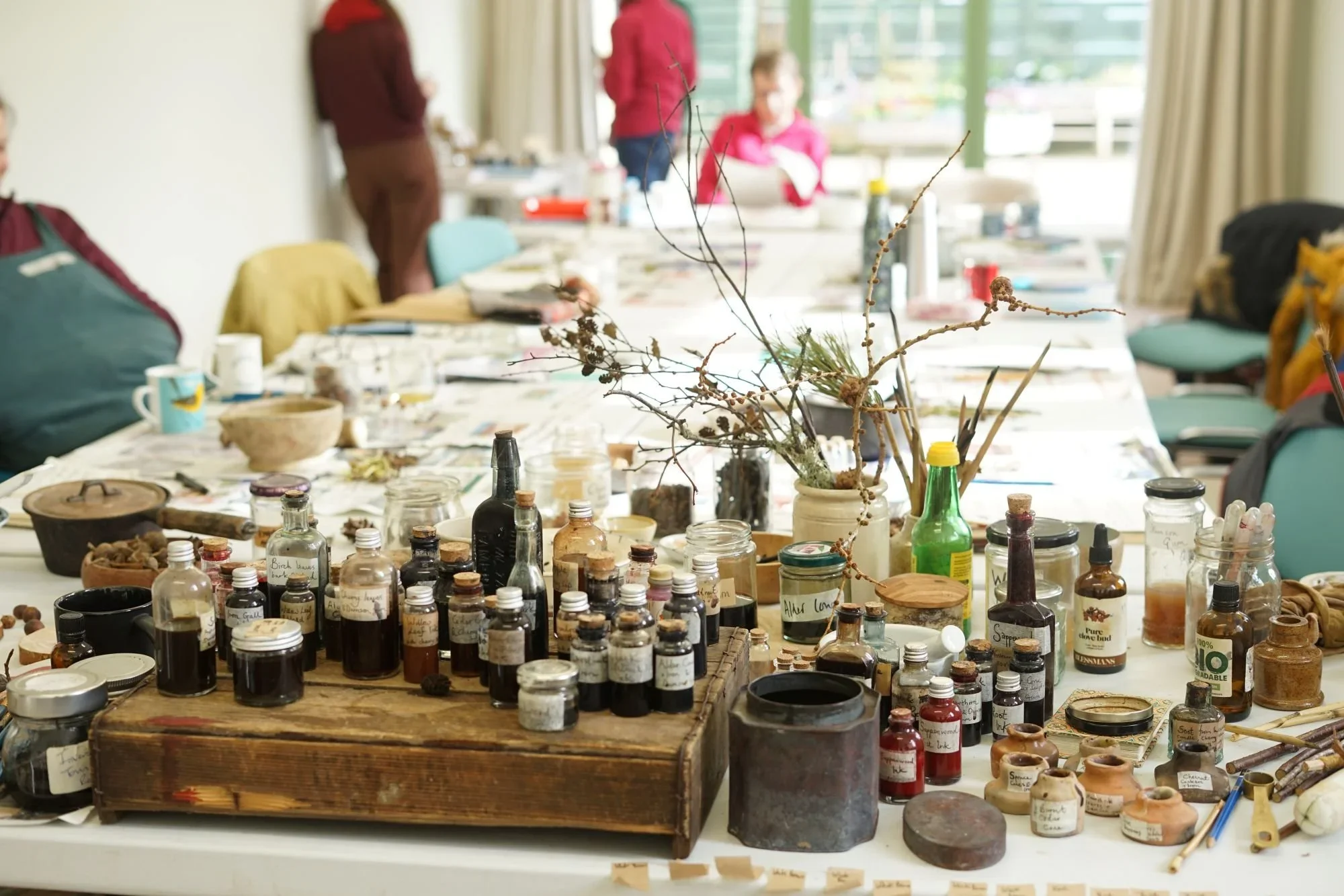 A table filled with various bottles, jars, and containers labeled with handwritten notes, and a vase with dried branches, in a well-lit room with people in the background.