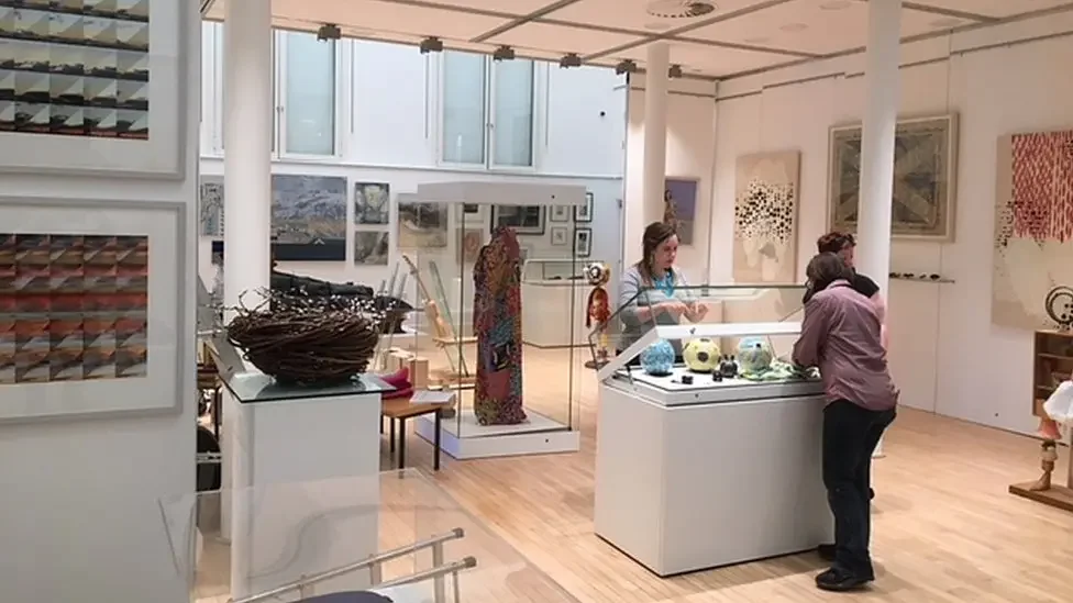 People observing art and glassware displays in an art gallery with paintings on the walls.