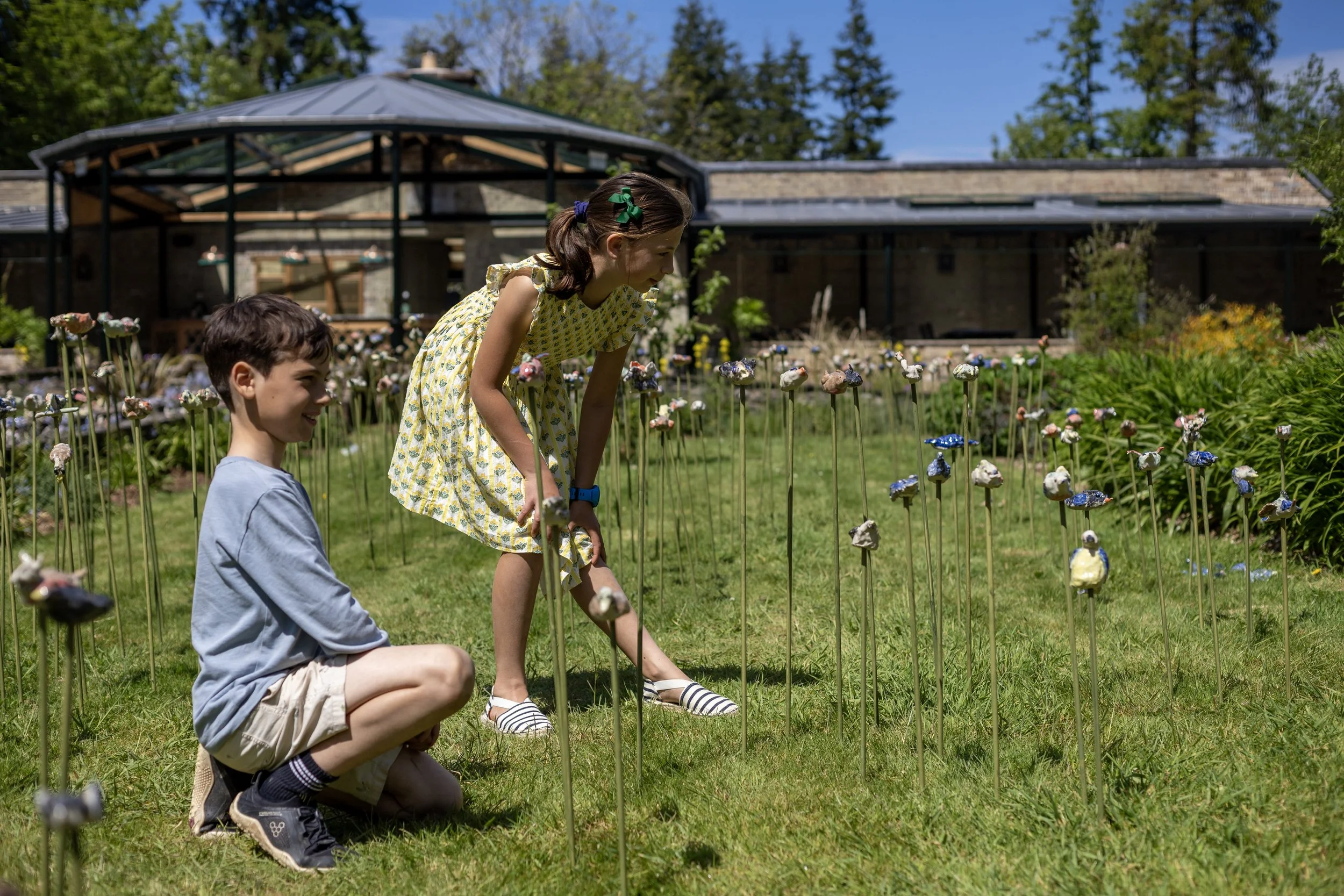 Two children, a girl in a yellow dress and a boy in a light blue shirt, are looking at and touching decorative mushrooms on sticks in a garden. There is a building with a patio and trees in the background.