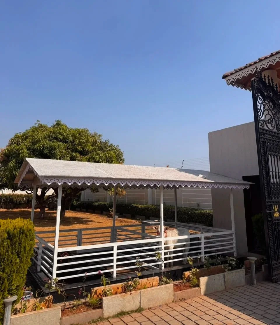 A small white wooden house with a sloped roof, decorated with intricate cutouts along the edges, enclosed by a white railing, with a goat standing inside. There are plants and a brick pathway outside, and a large green tree in the background under a clear blue sky.