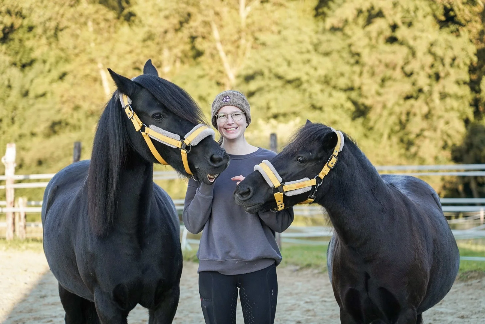 Junge Frau mit Brille und Mütze steht zwischen zwei schwarzen Ponys auf einem Reitplatz, umgeben von einem Baumhintergrund.