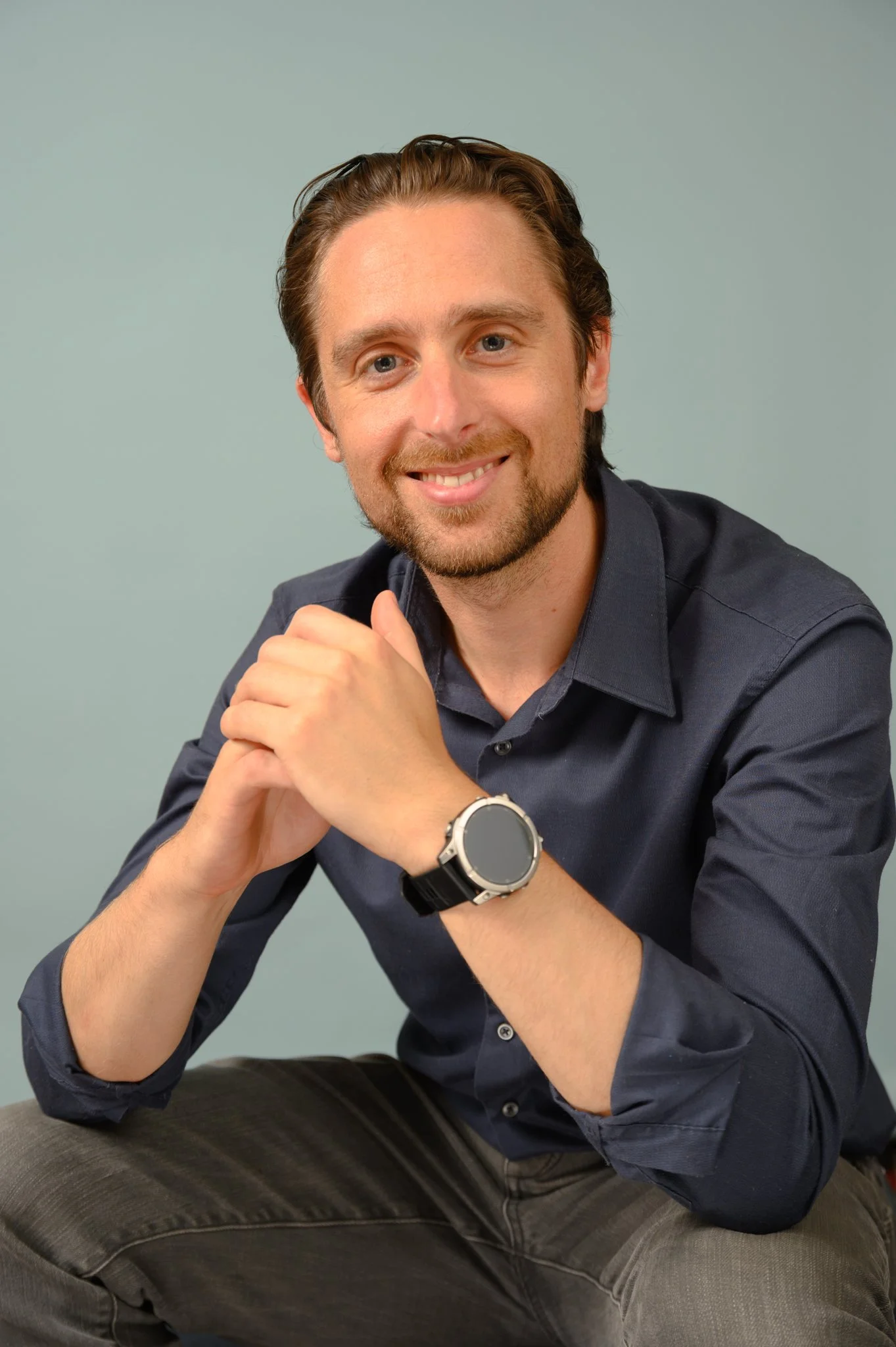 Dr Jake Maxwell Watts is a man with brown hair, beard, and blue eyes is sitting and smiling at the camera. He wears a dark button-up shirt. His hands are clasped together, resting on his knee, against a muted green background.