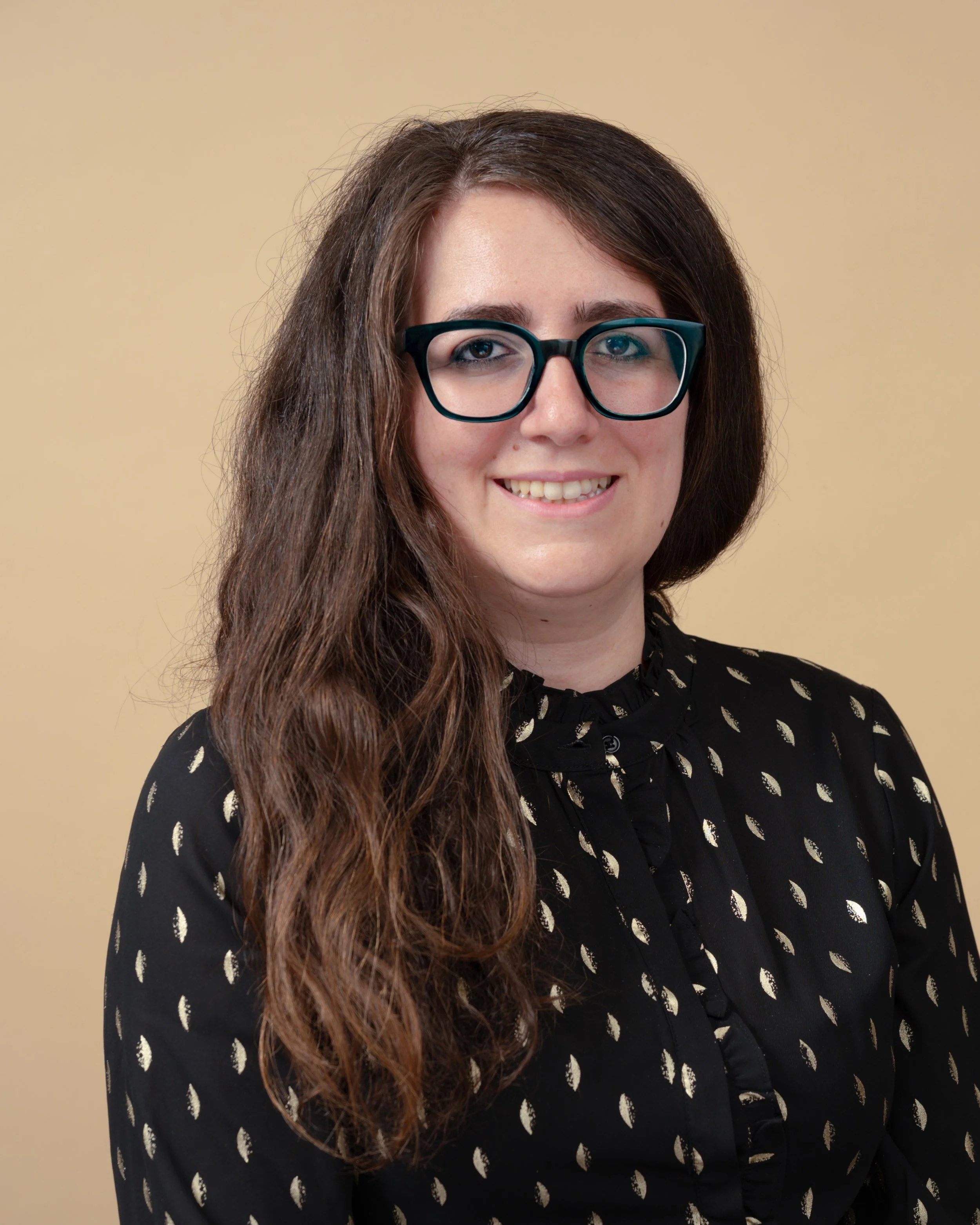 Dr Olivia Mohtady is a woman with long, wavy brown hair wearing black glasses and a black blouse with white feather patterns, smiling against a beige background.