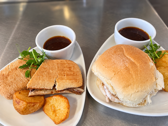 Two plates of pulled pork sandwiches with dipping sauce, greens, and fried potato sides