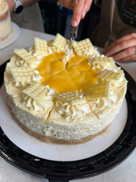 A person slicing into a creamy white cake topped with white chocolate pieces and a caramel center, on a black cake stand.
