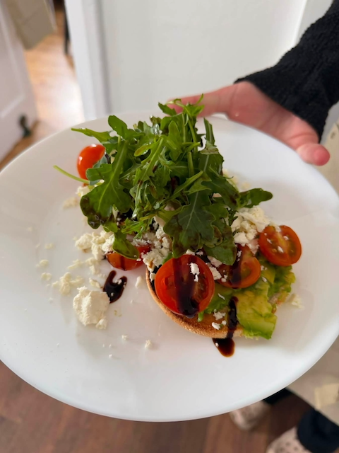 A plate with a salad that includes cherry tomatoes, leafy greens, crumbled cheese, and balsamic glaze on a slice of toasted bread.
