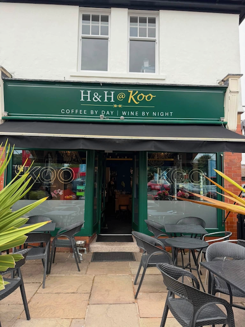 Exterior view of a coffee shop named H&H @ Koo with outdoor seating, black tables and chairs, green storefront and signage, large windows, situated on a street with plants on either side.