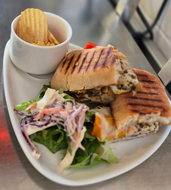 Grilled sandwich with coleslaw and leafy salad on a white plate, served with ridged potato chips in a cup