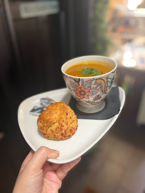 Hand holding a white tray with a floral teacup of orange soup garnished with herbs and a savory cheese scone.