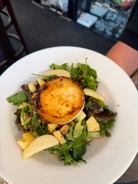 Salad with a baked or grilled scallop on top, mixed greens, apple slices, and cheese on a white plate.