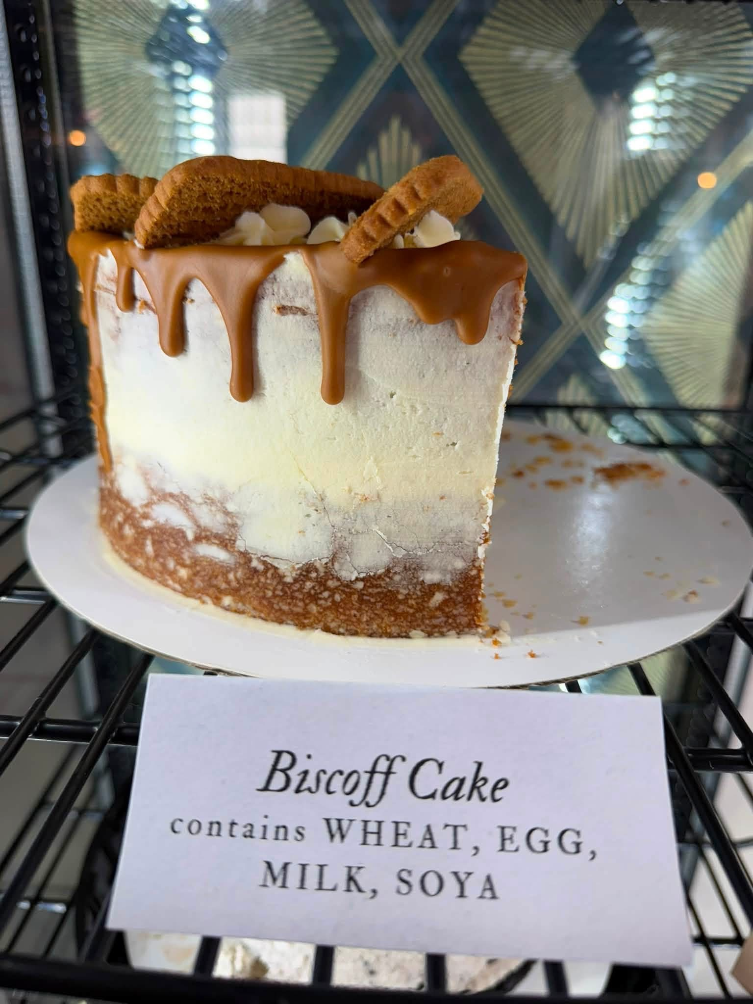 Close-up of a slice of biscuit cake with caramel drizzle, topped with two biscuit cookies and some white cream decoration, on a white plate. There is a sign underneath with text partially visible, stating 'Biscuit Cake' and listing ingredients like wheat, eggs, milk, and soya.