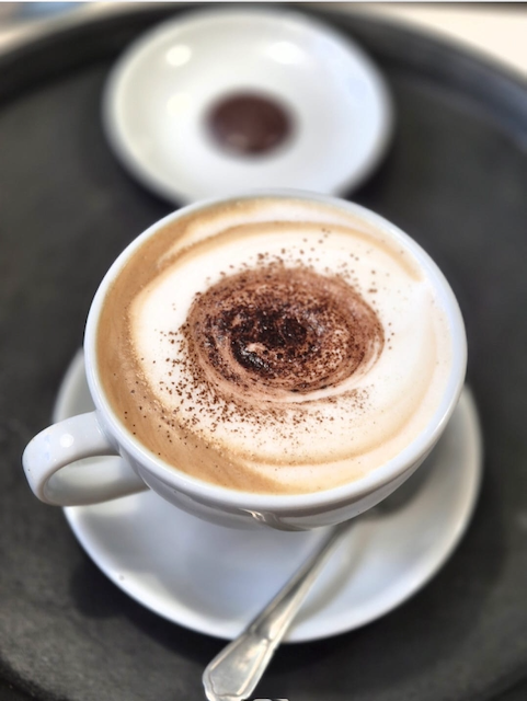 White cup of cappuccino topped with cocoa powder on a saucer with a spoon, served on a black tray