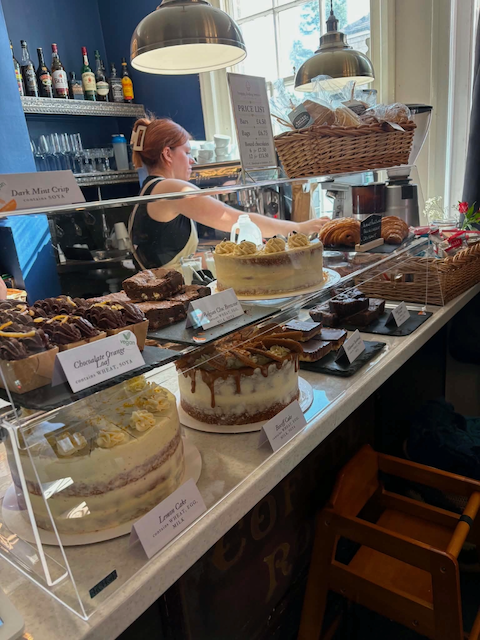 A woman working behind a bakery counter displaying various cakes and pastries, with a basket of bread and bread rolls, and bottles and glasses on shelves in the background.