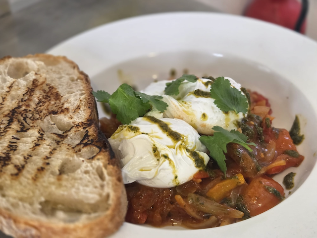 Toasted bread served with poached eggs, tomato and pepper sauce, cilantro, and herb oil in a white bowl