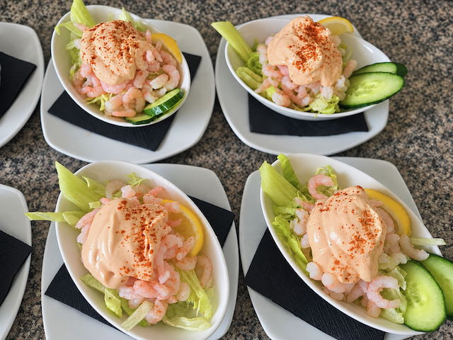 Four bowls of shrimp salad with lettuce, cucumber, lemon, and creamy paprika-topped dressing on white plates.