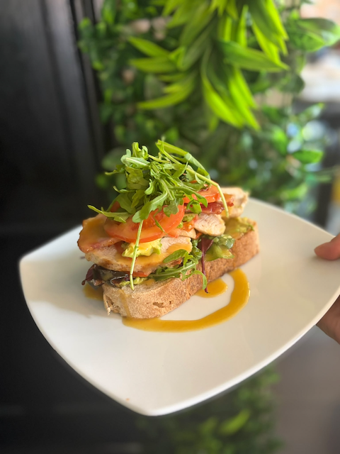 Gourmet open-faced sandwich with toasted bread, eggs, tomatoes, greens, avocado, and microgreens on a white plate with sauce drizzle