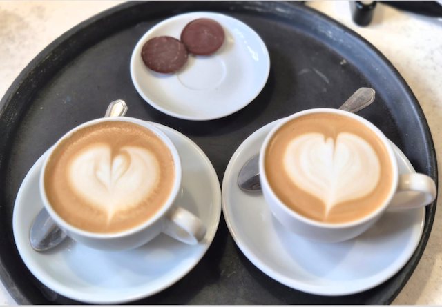 Two cups of latte art coffee on saucers with spoons and two chocolate cookies on a black serving tray