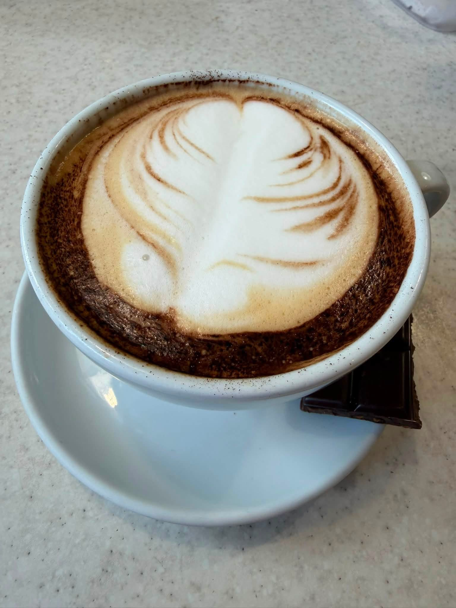 A cup of coffee with frothy milk and cinnamon or cocoa powder design on top, sitting on a white saucer on a beige countertop.