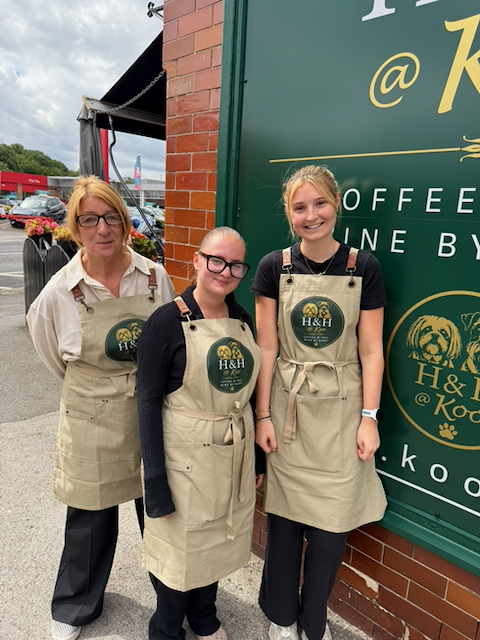 Three women standing outside a coffee shop, all wearing aprons with the H&H logo. Two are smiling and standing close together, while the woman on the left has a neutral expression. The background shows a brick wall and a large green sign.