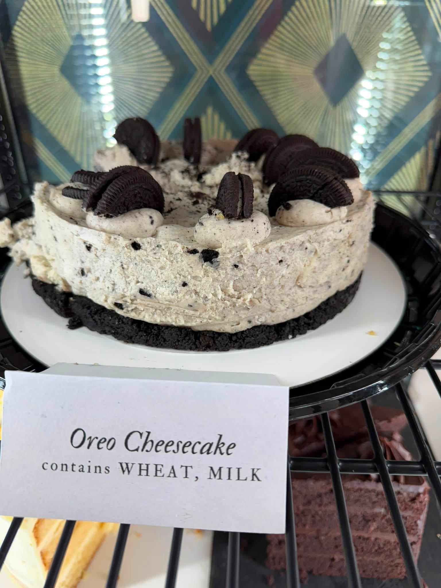 Oreo Cheesecake on display, topped with Oreo cookies, with a label indicating it contains wheat, eggs, milk, and other ingredients, in a bakery or cafe setting.