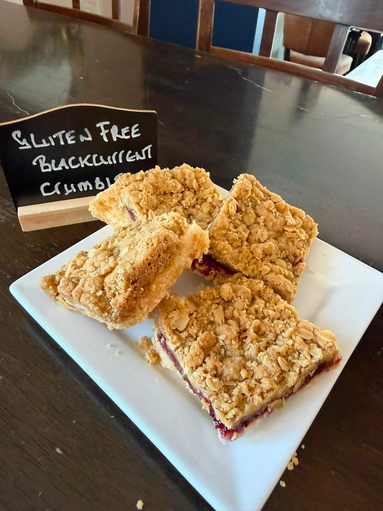 Rectangular crumbly dessert bars with a berry filling on a white plate, next to a blackboard sign that reads 'Gluten Free Blackberry Crumble'.