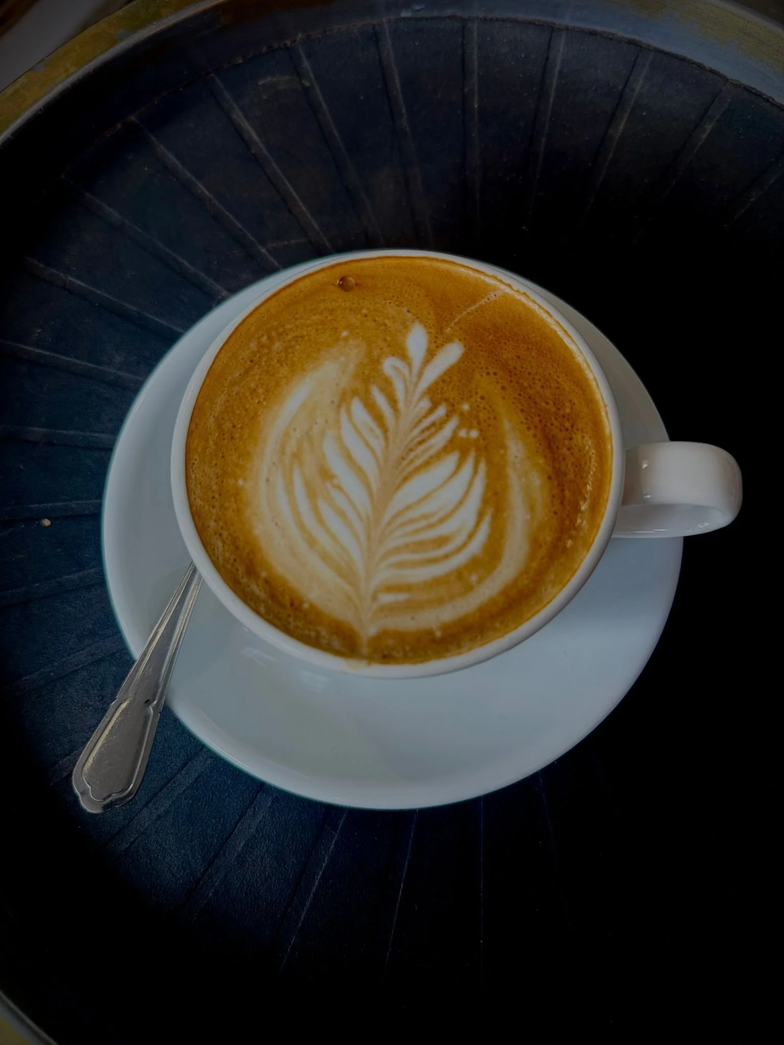 A cup of latte with latte art on top, placed on a saucer with a spoon, inside a dark coffee shop.