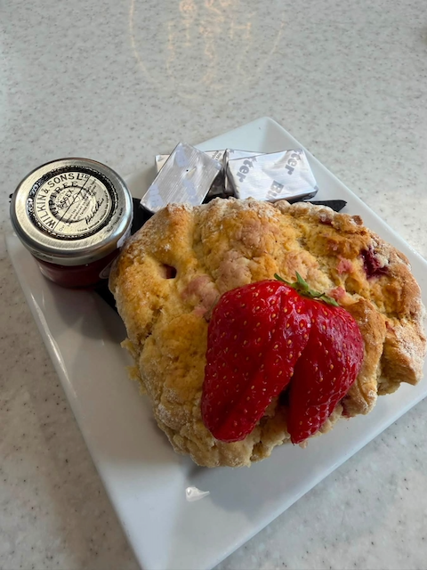 Fried chicken with a large strawberry on top, a small jar of jam, and a packet of butter on a white square plate, placed on a light-colored countertop.