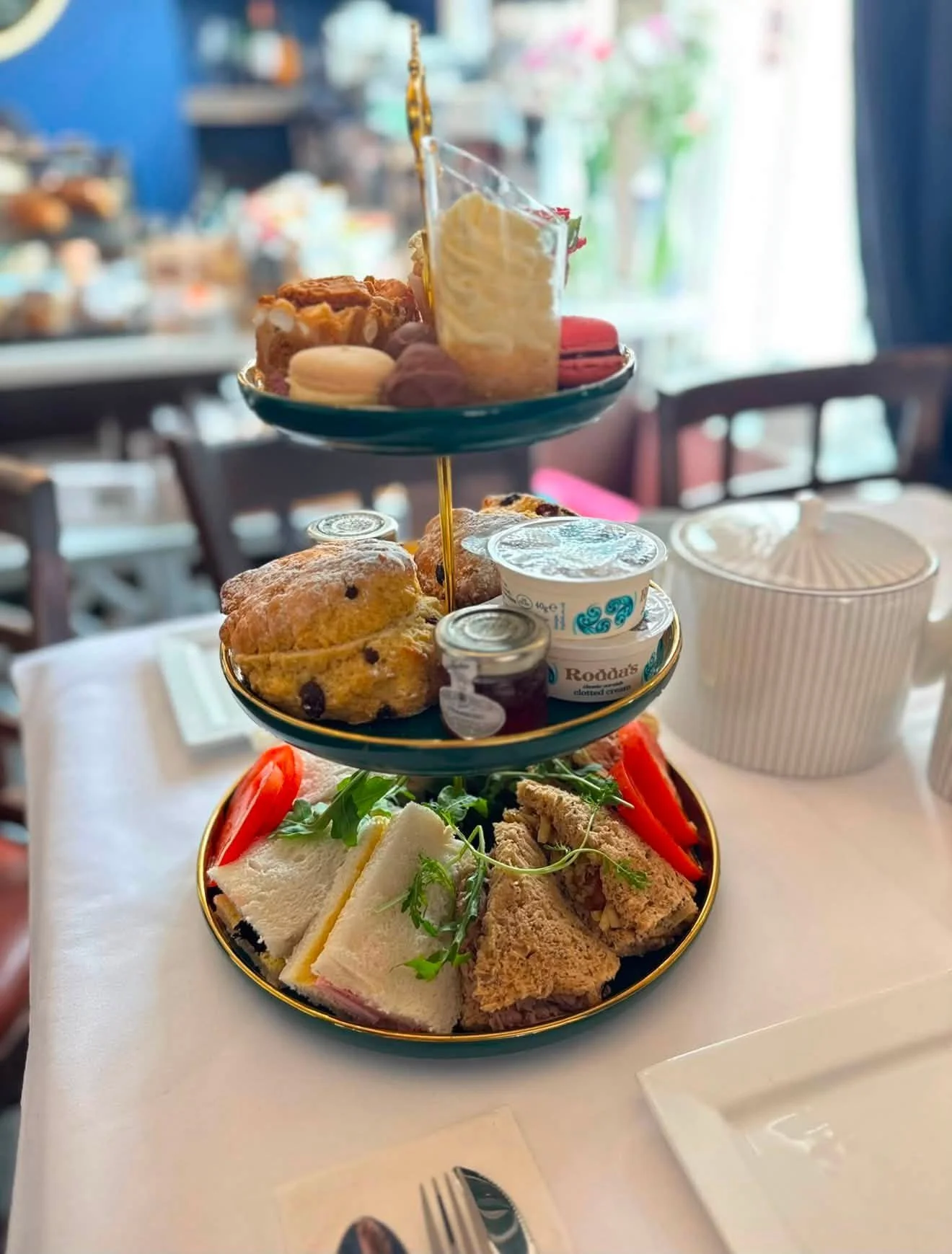 Three-tiered serving tray holding assorted cookies, macarons, finger sandwiches, cheeses, and salads at a restaurant