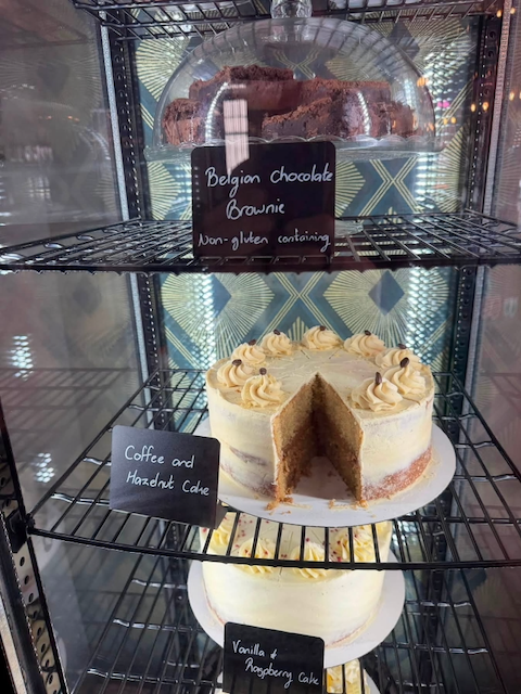 Display case with baked goods including a gluten-free Belgian chocolate brownie on the top shelf, a coffee and hazelnut cake with a slice removed on the middle shelf, and a vanilla and raspberry cake on the bottom shelf.