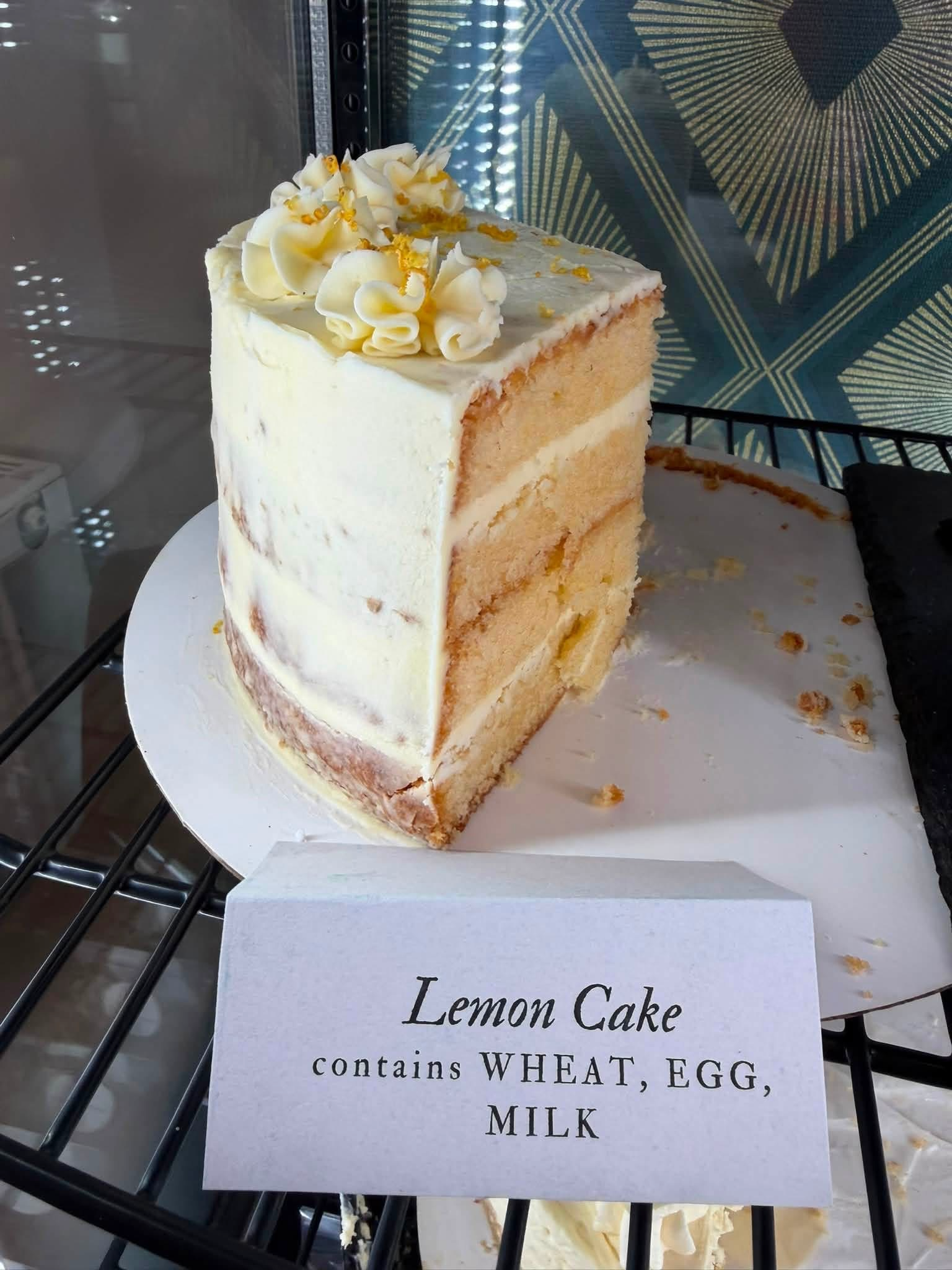 A slice of lemon cake with white frosting and decorative white frosting on top, displayed in a bakery case with a label reading "Lemon Cake" containing wheat, egg, and milk.