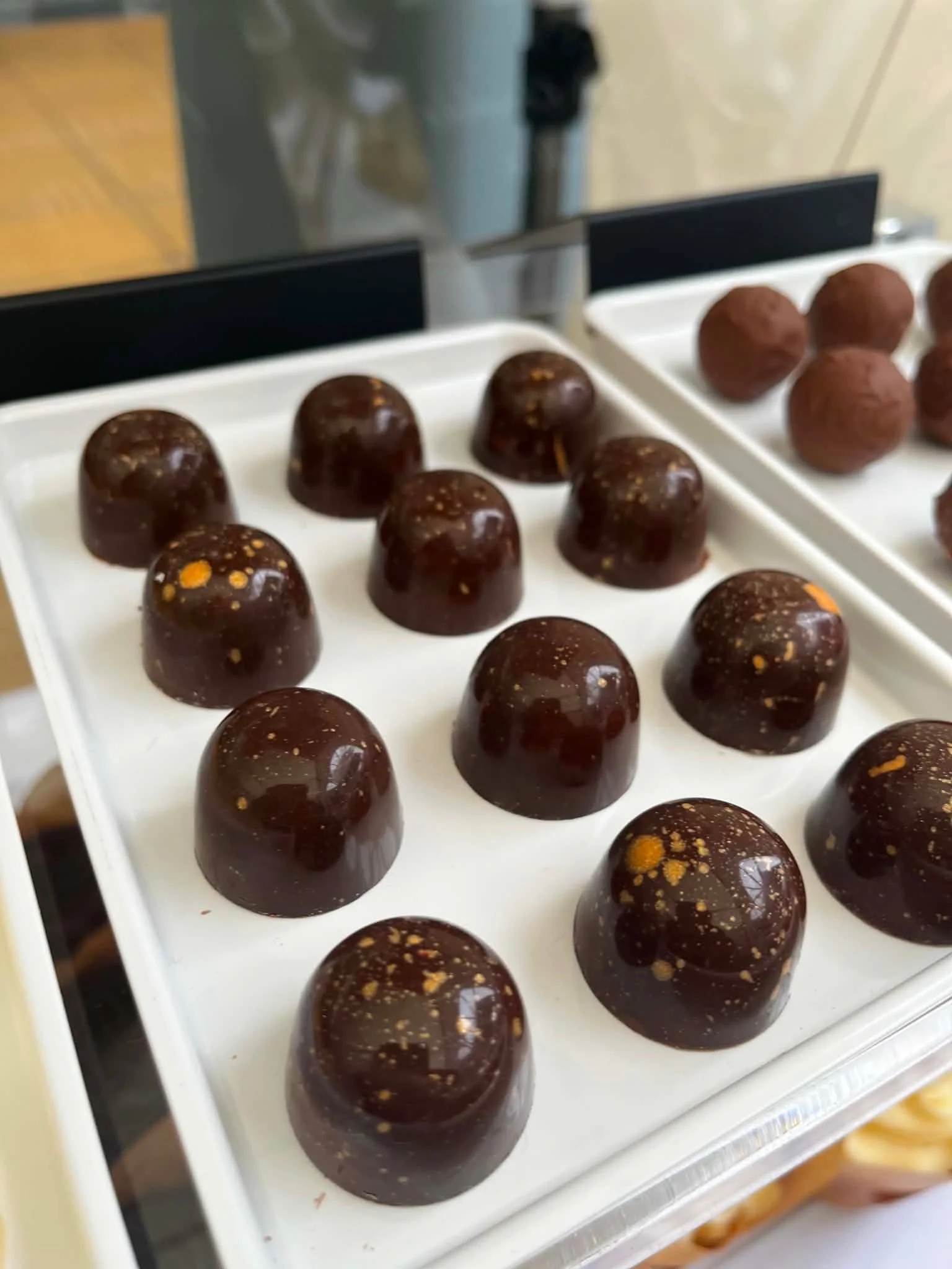 Close-up of tray filled with round chocolate truffles with gold specks, on a white tray with black dividers, in a kitchen setting.