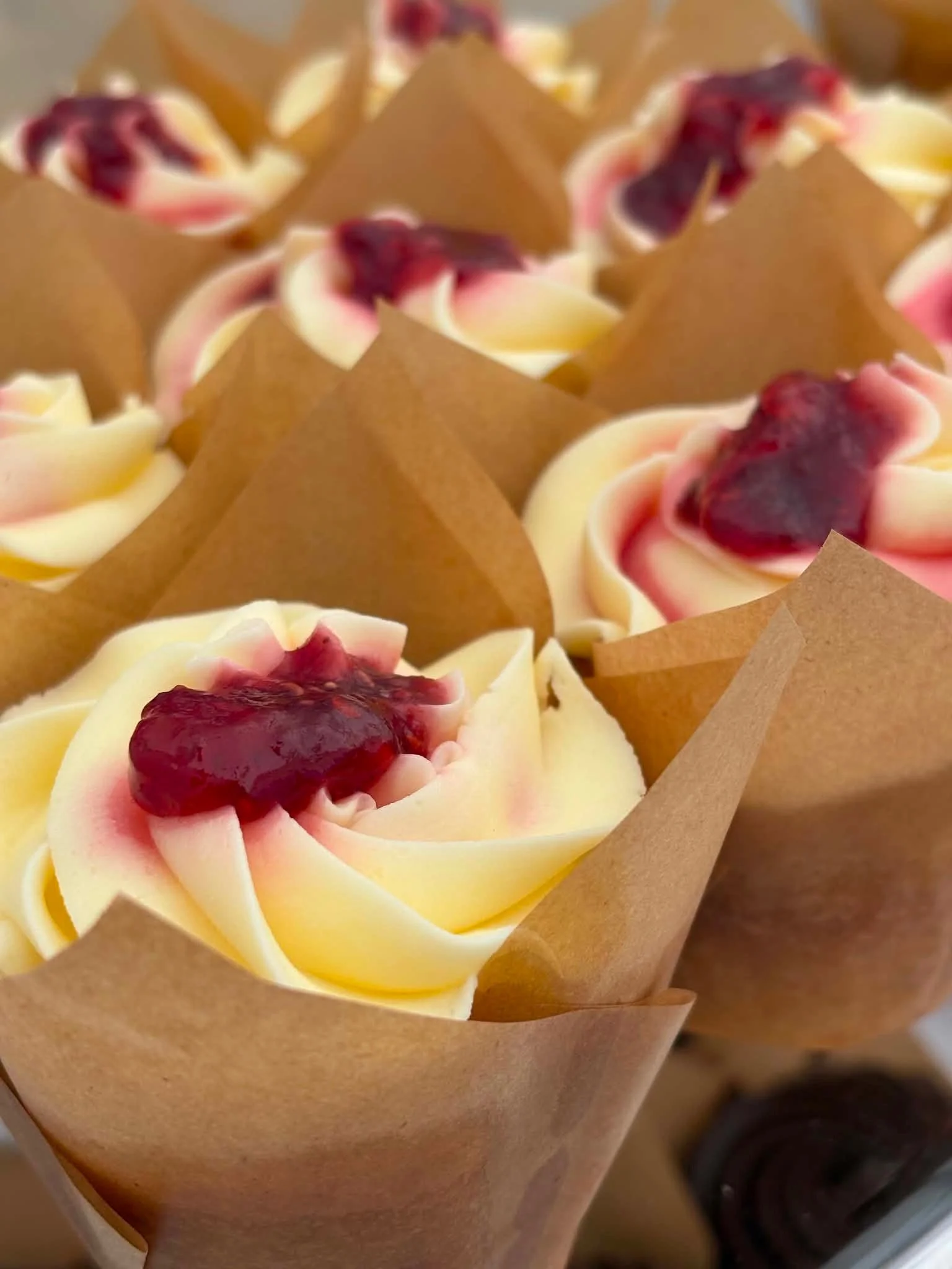 Cupcakes with white frosting, topped with a dollop of red fruit jam, wrapped in brown parchment paper.