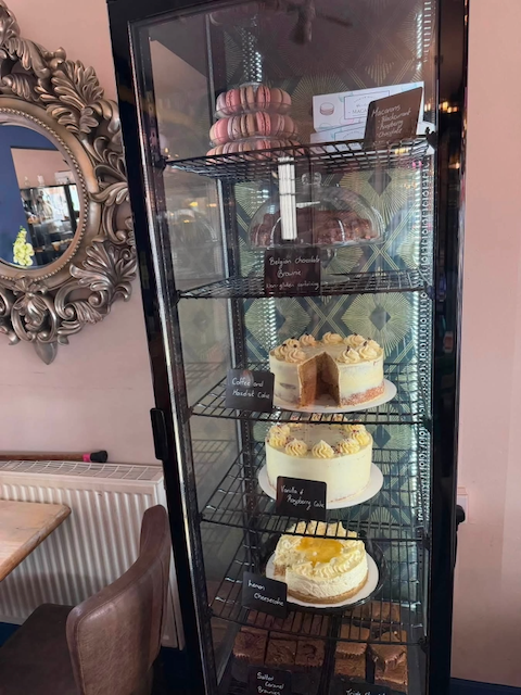 A glass display refrigerator containing macarons, salted caramel cake, vanilla sponge cake, and honey cheesecake on glass shelves in a cafe or bakery.