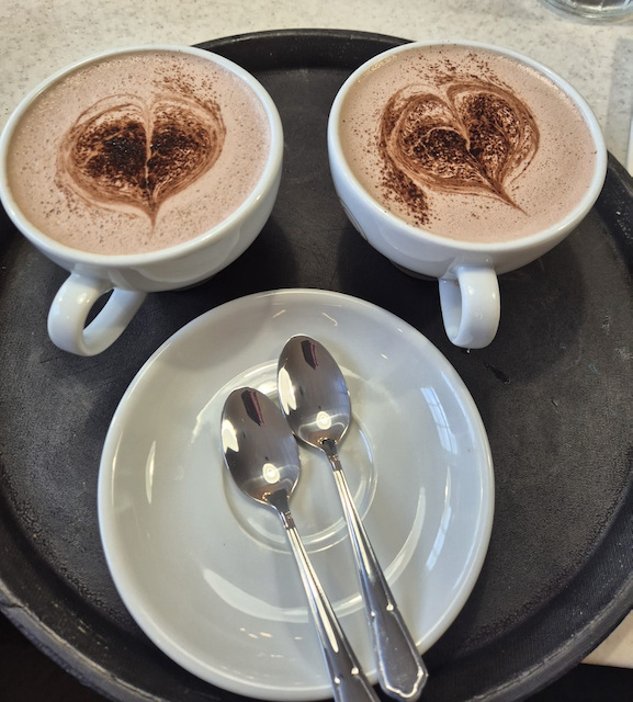 Two cups of cappuccino with heart-shaped cocoa designs on a black café tray, accompanied by a white saucer and two teaspoons.