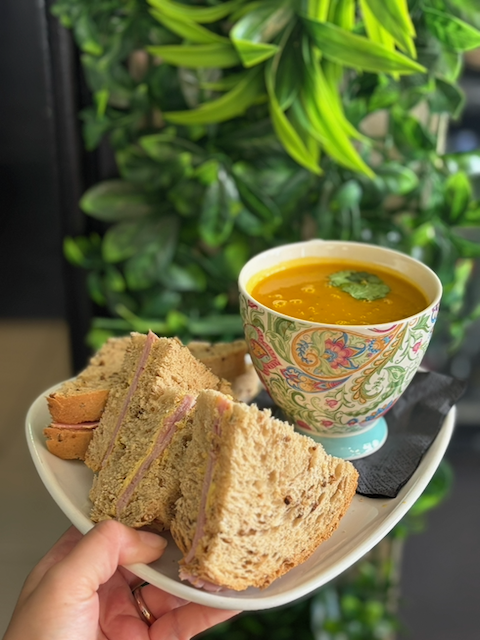 Plate of triangular tea sandwiches with pink filling beside a floral teacup of orange soup, garnished with cilantro