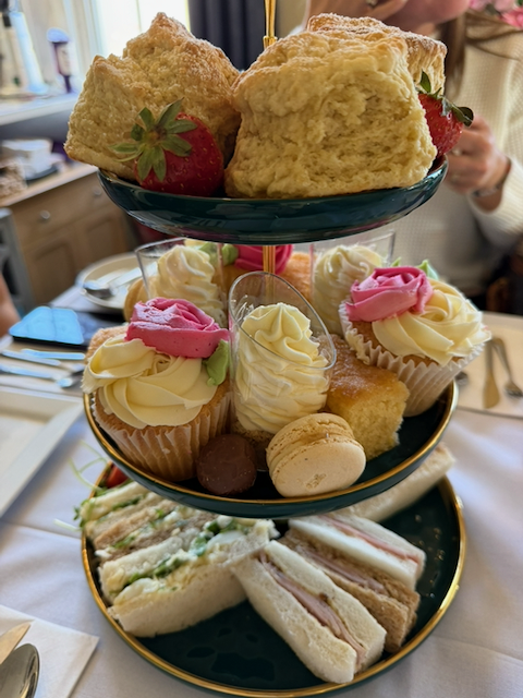 Three-tier afternoon tea stand with scones, strawberries, frosted cupcakes, macarons, pastries, and assorted finger sandwiches.
