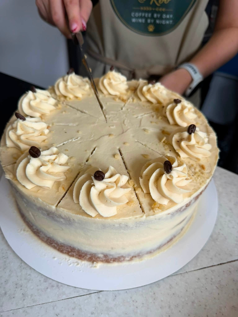 A person cutting a round vanilla cake with whipped cream and coffee bean decorations on top.