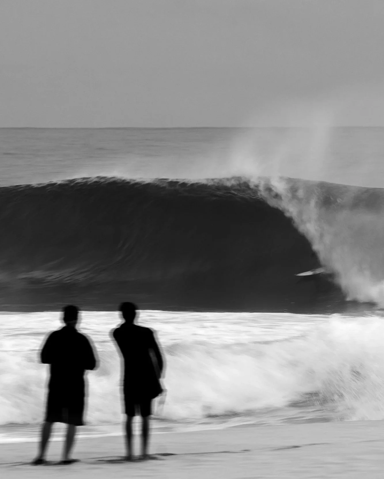 Two people stand on the beach watching a large ocean wave with a surfer riding inside it, captured in black and white.