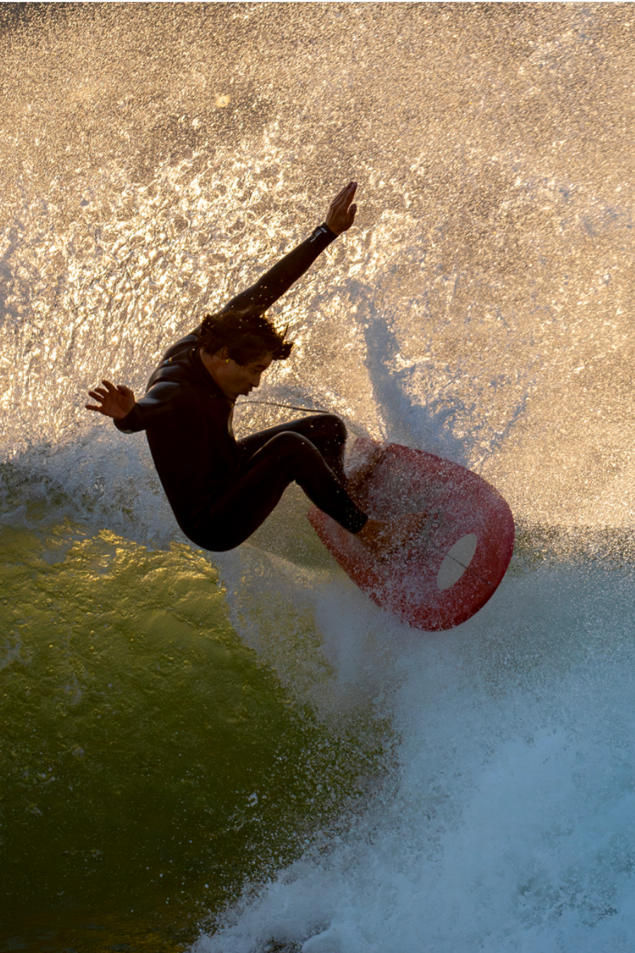 Person wakeboarding on a lake during sunset, wearing a black wetsuit, with water splashing around.