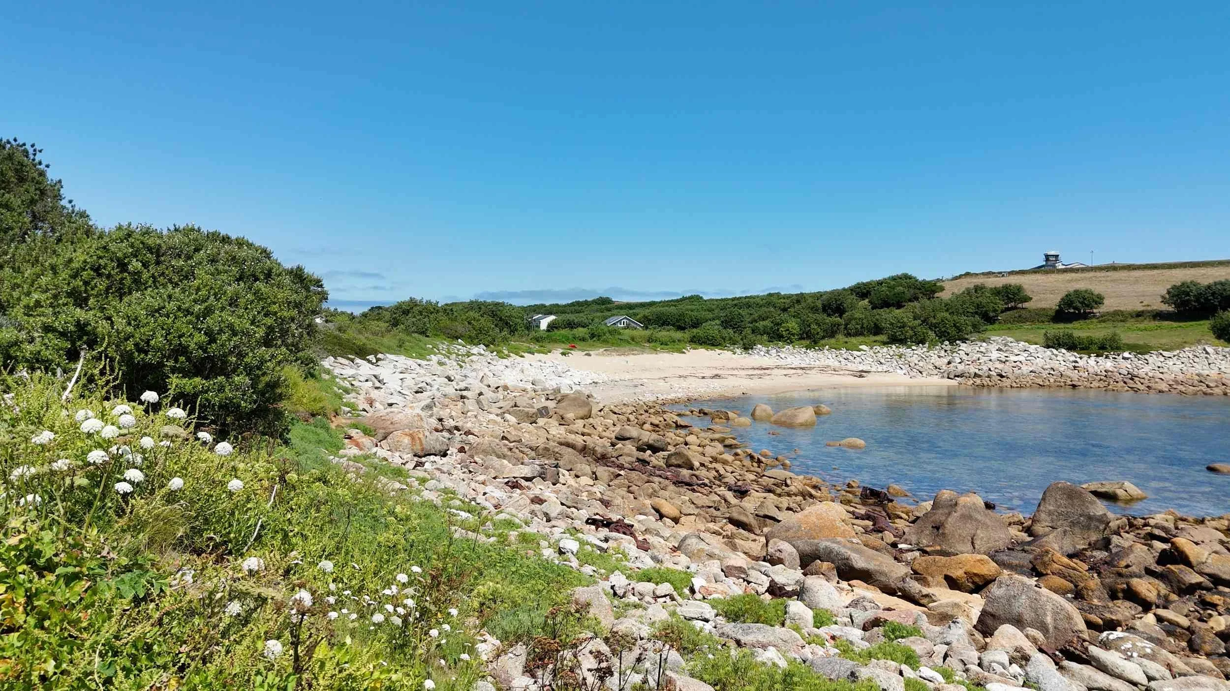 A rocky beach with large stones along the shoreline, green bushes, and grass. There are a few distant small houses and a small airport on the hilltop under a clear blue sky.