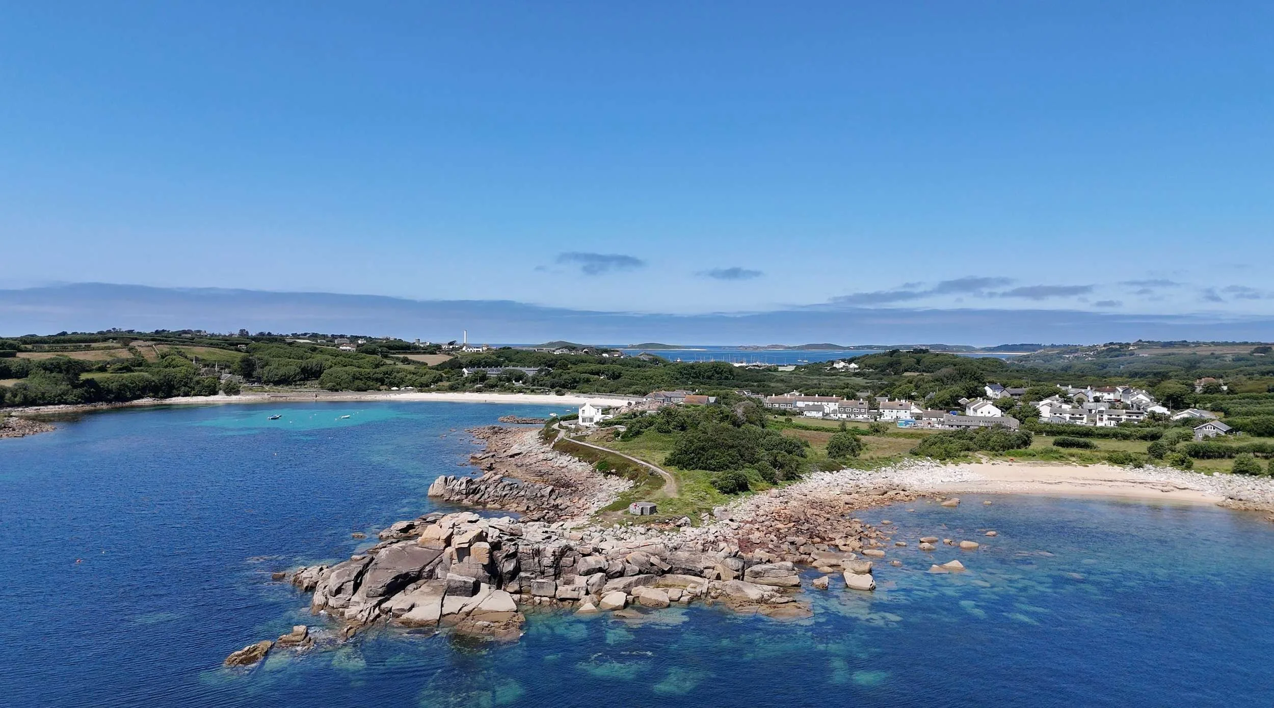 Scenic coastal landscape with two calm blue bays, rocky shoreline, white houses, green trees, and distant islands beyond, all under a clear blue sky.