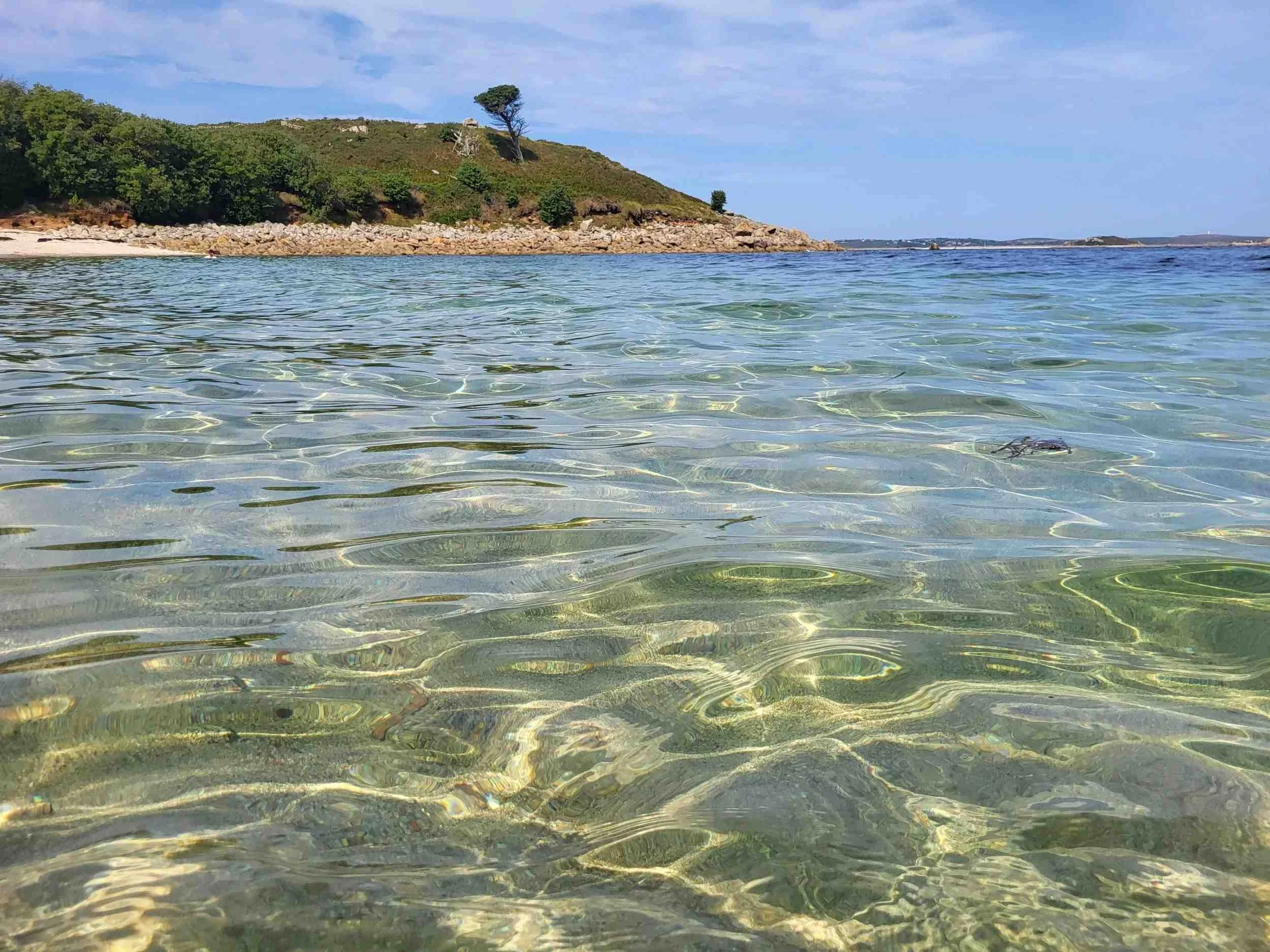 Clear ocean water with a rocky shoreline and a green, hill in the background under a blue sky with scattered clouds.