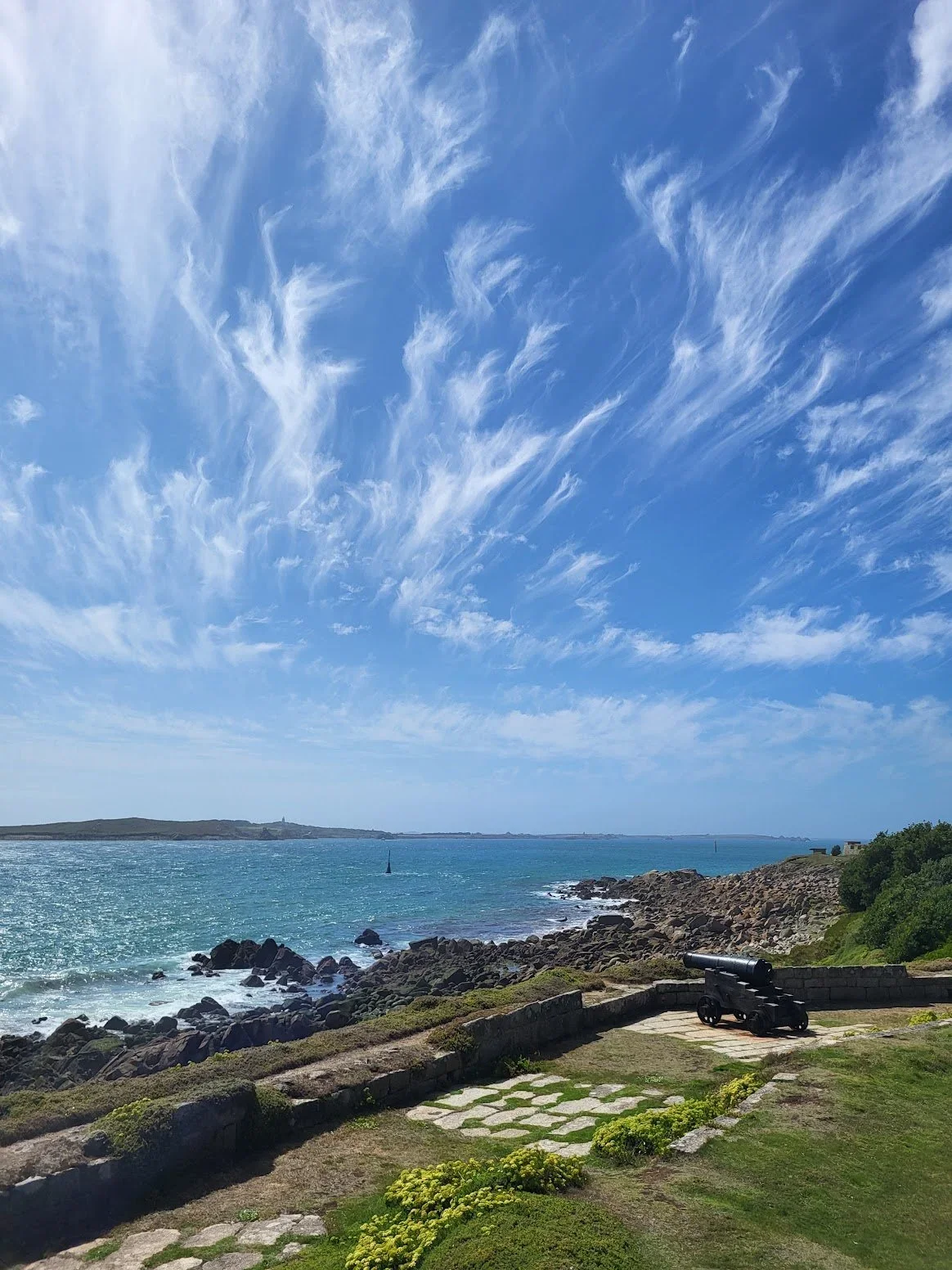 A scenic coastal view with blue sky and wispy clouds, rocky shoreline, turquoise water, and a cannon on a stone platform in the foreground.