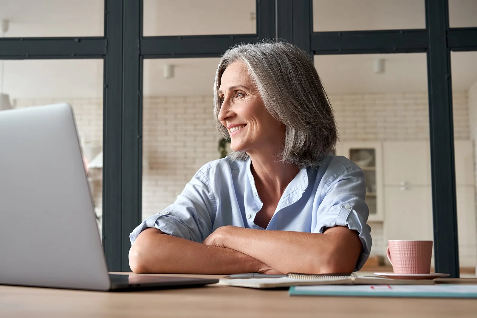 Older woman with gray hair working on a laptop at a desk, smiling, in a modern home office with a glass partition and kitchen in the background.