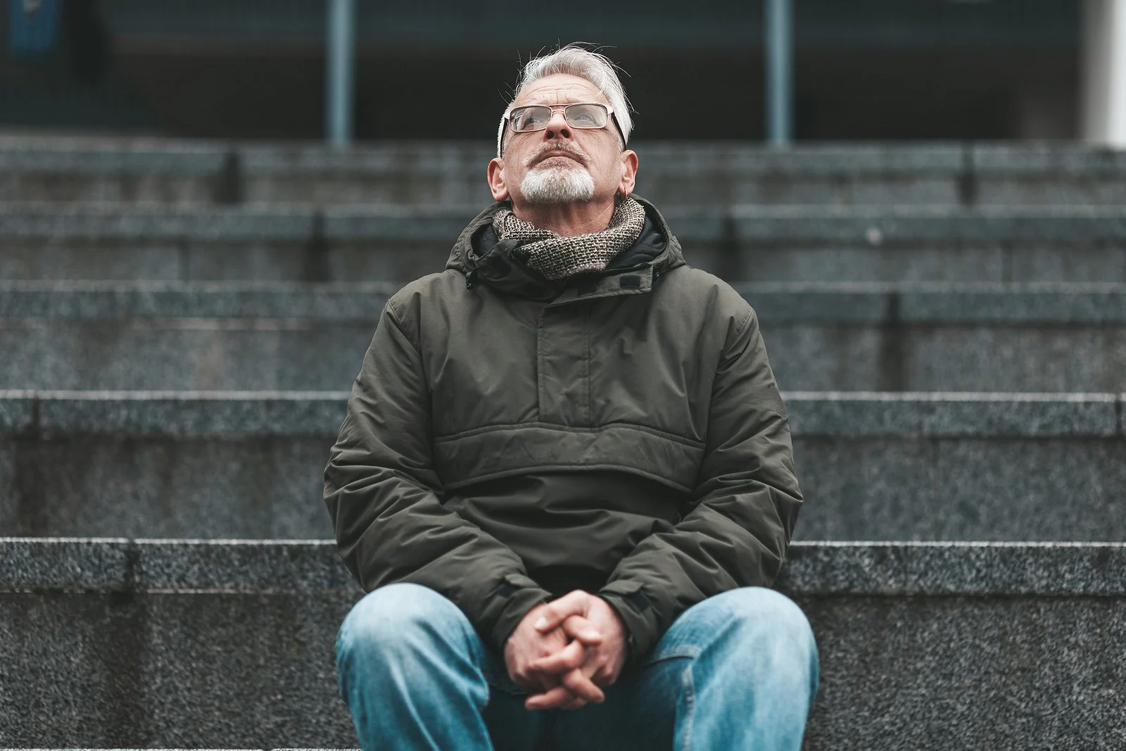 An elderly man with gray hair and a beard, wearing glasses, sitting on gray stone steps outdoors, looking upward, reflecting on his future.