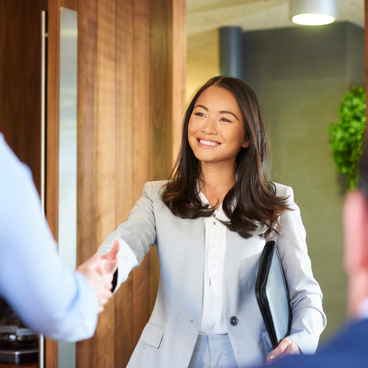 Businesswoman in gray suit smiling and shaking hands with a colleague in a modern, professional office setting.