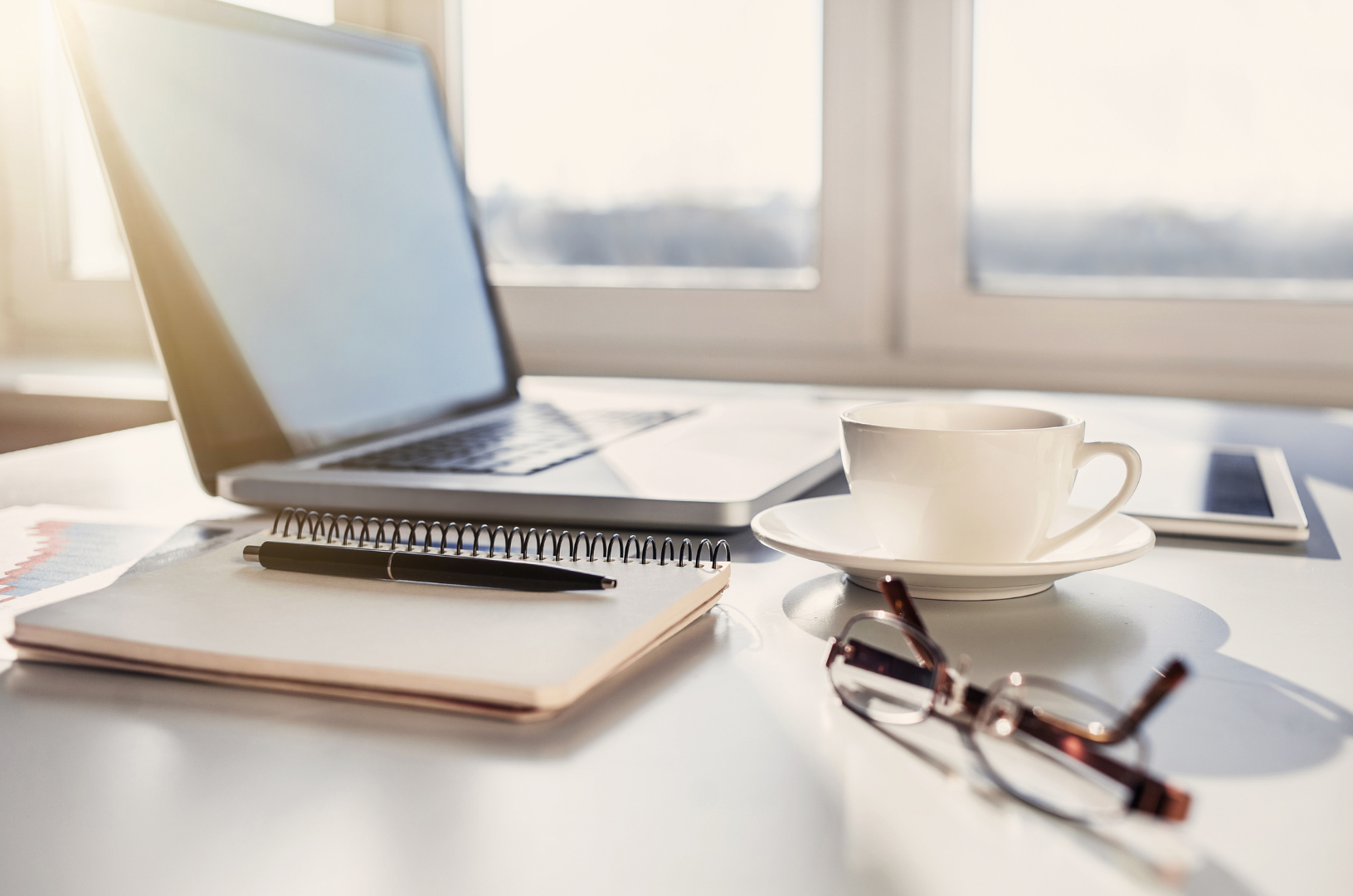 A workspace with a laptop, a spiral notebook with a pen, a pair of eyeglasses, a white cup on a saucer, a smartphone, and a calendar or planner on a white table, with sunlight streaming through a window in the background.