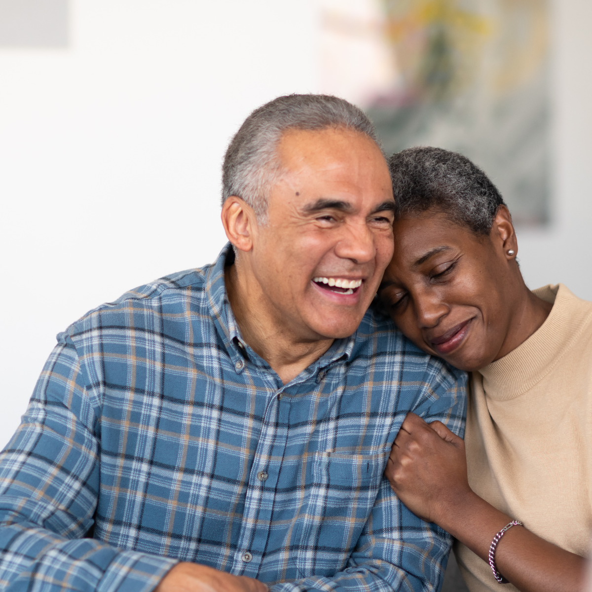 An older man and woman sharing a happy, intimate moment, with the woman leaning her head on the man's shoulder and both smiling.