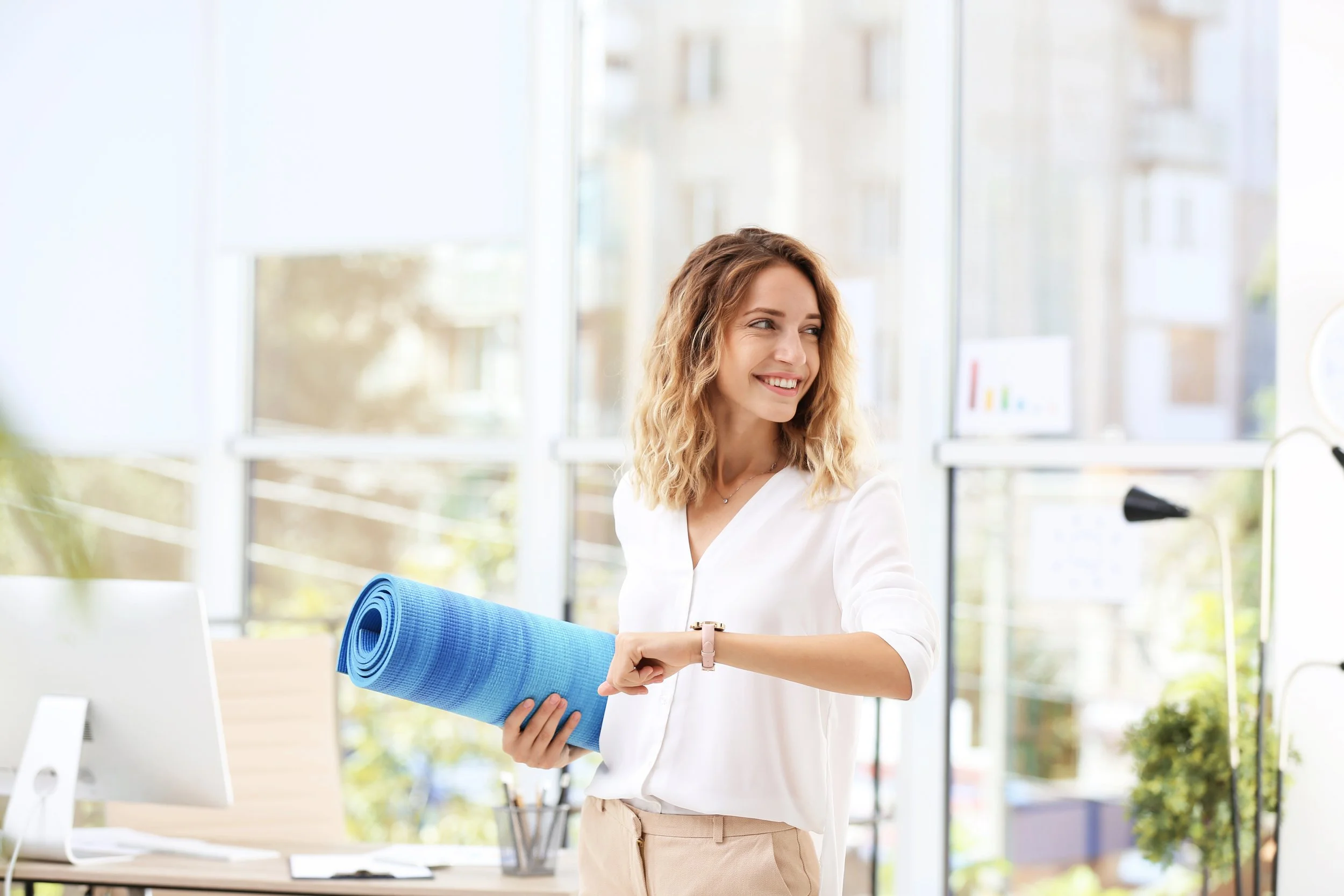 A woman smiling and looking to her left holding a blue yoga mat and a clipboard inside a bright, modern office or studio with large windows.