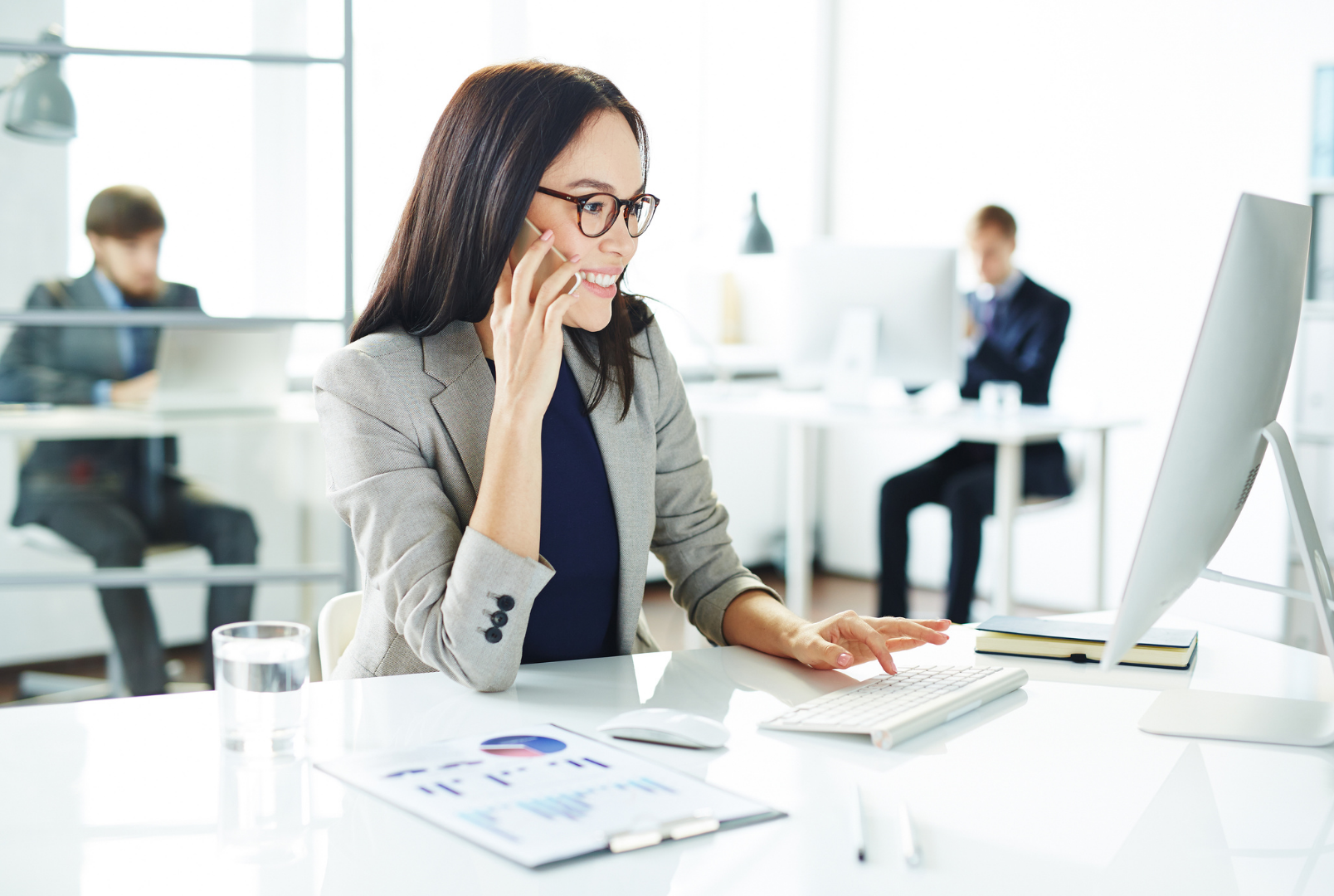 Businesswoman sitting at a desk, talking on her cellphone, working on a computer in a modern office with other employees working at desks in the background.